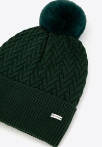 Ciemnozielona dzianinowa czapka z teksturowanym wzorem zygzakowatym i puszystym, turkusowym pomponem na szczycie; posiada prążkowany mankiet i małą metkę.