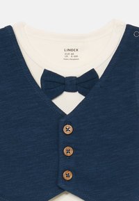 Weißer Baby-Body mit einem vorn angenähten marineblauen Weste, drei Holzknöpfen und einer passenden marineblauen Stofffliege.