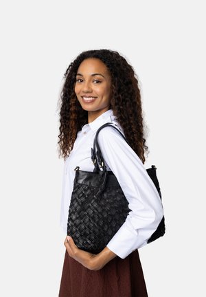 Glunderende vrouw met krullend haar, gekleed in een witte blouse en een bruine rok, houdt een zwarte geweven leren handtas over haar schouder tegen een effen achtergrond.