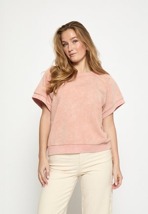 Lichtroze sweatshirt met korte mouwen, een ontspannen pasvorm en geribbelde zoom. Zachte textuur met een licht verwassen afwerking. Draagt beige broek.