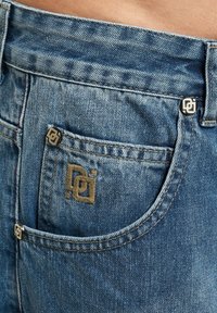 Lys blå denimjeans med en falmet tekstur. Har et gullbrodert logo og metallakenter på lommen og livet.