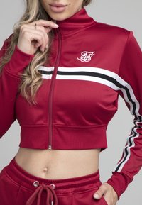 Rød cropped zip-up jakke med hvide og sorte striber på ærmerne og et lille logo på brystet, lavet af et glat, strækbart materiale.