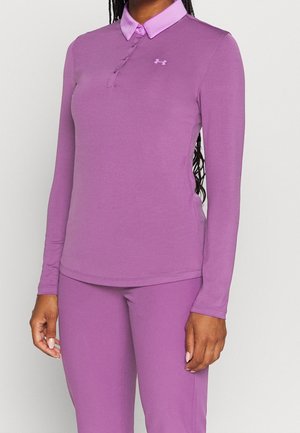 Kobieta w dopasowanej liliowej koszulce polo z długim rękawem i patką na guziki oraz dopasowanych spodniach, prezentująca styl ubrań sportowych.