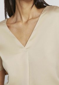 Beige blus med v-ringning, korta ärmar, lätt material och en mjuk textur med minimal sömnadsdetaljer.
