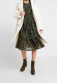 Un manteau en fausse fourrure blanche sur une robe noire à motif floral avec un ourlet évasé. Porté avec des bottines à talons vertes présentant un motif texturé.