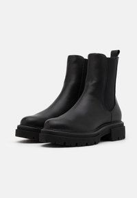 Une paire de bottines en cuir noir avec des semelles en caoutchouc épaisses et des panneaux latéraux en élastique, conçues pour un enfilage facile.