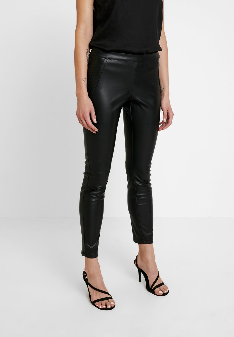 Femme portant un haut noir sans manches, un pantalon slim en cuir noir et des sandales noires à talons hauts sur fond blanc.