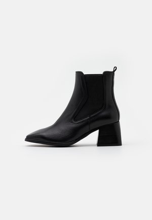 Bottine en cuir noir avec bout pointu, talon bloc, panneaux latéraux élastiques et languette de traction sur fond blanc uni.
