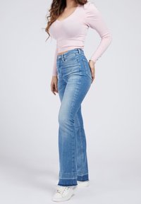 Jeans évasés bleu clair à taille haute, avec un ourlet brut, un design à cinq poches et un délavage. Associés à un haut ajusté rose et des baskets blanches.
