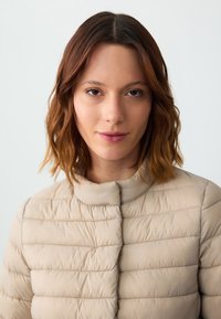 Giovane donna con capelli castani di lunghezza media, che indossa una giacca trapuntata beige, rivolta in avanti su uno sfondo chiaro uniforme.