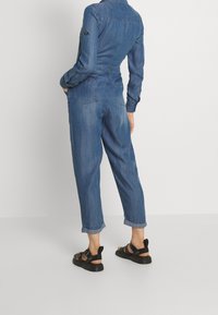 Mono de mezclilla con mangas largas, bolsillos traseros y puños enrollados. Presenta un diseño ajustado en azul medio. Combinado con sandalias negras.