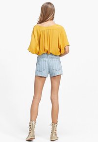 Top amarillo, holgado, con la parte trasera fruncida, combinado con pantalones cortos de mezclilla de tiro alto de un azul claro con el dobladillo deshilachado. Botas de tobillo de encaje beige.