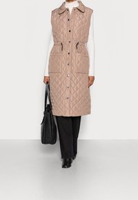 Gilet matelassé beige avec un col, boutons-pression à l'avant, taille à cordon de serrage et deux poches latérales, associé à un pantalon noir et un sac.