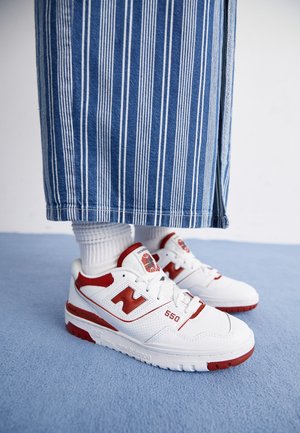 Zapatillas New Balance 550 blancas y rojas, usadas con calcetines blancos y pantalones a rayas azules y blancas sobre una alfombra azul.
