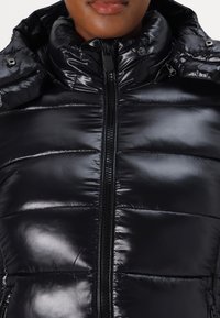 Gros plan sur une personne portant une doudoune noire brillante avec un col montant zippé et des détails à boutons-pression.