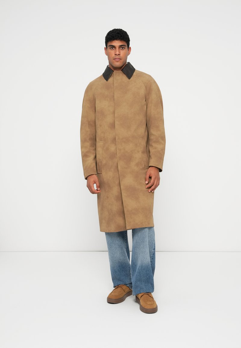 Long tan coat with a dark collar, front pockets, smooth texture, paired with blue wide-leg jeans and brown suede shoes.