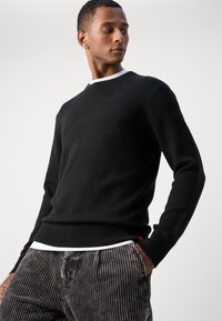 Suéter de punto negro con cuello redondo y dobladillo acanalado, superpuesto a una camisa blanca. Usado con pantalones oscuros de rayas, que muestran un corte relajado.