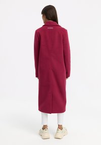 Fleecejacka i djup rosa med sned krage och rak passform. Har en slits i ryggen och en etikett i nacken. Bärs med vita byxor och sneakers.