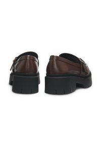 Chaussures en cuir marron avec une semelle en caoutchouc noire épaisse, dotées d'un talon surélevé et d'un détail de boucle sur le côté. Texture lisse, design minimaliste.