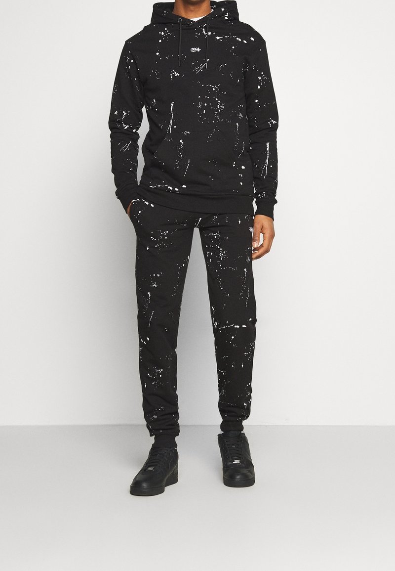 Homme portant un sweat à capuche noir et un pantalon de jogging avec un motif de gouttelettes de peinture blanche, ainsi que des baskets noires, debout devant un fond blanc uni.