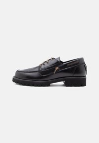 Chaussure en cuir noir avec un bout arrondi, quatre œillets marron, des coutures décoratives et une semelle en caoutchouc épaisse et texturée.