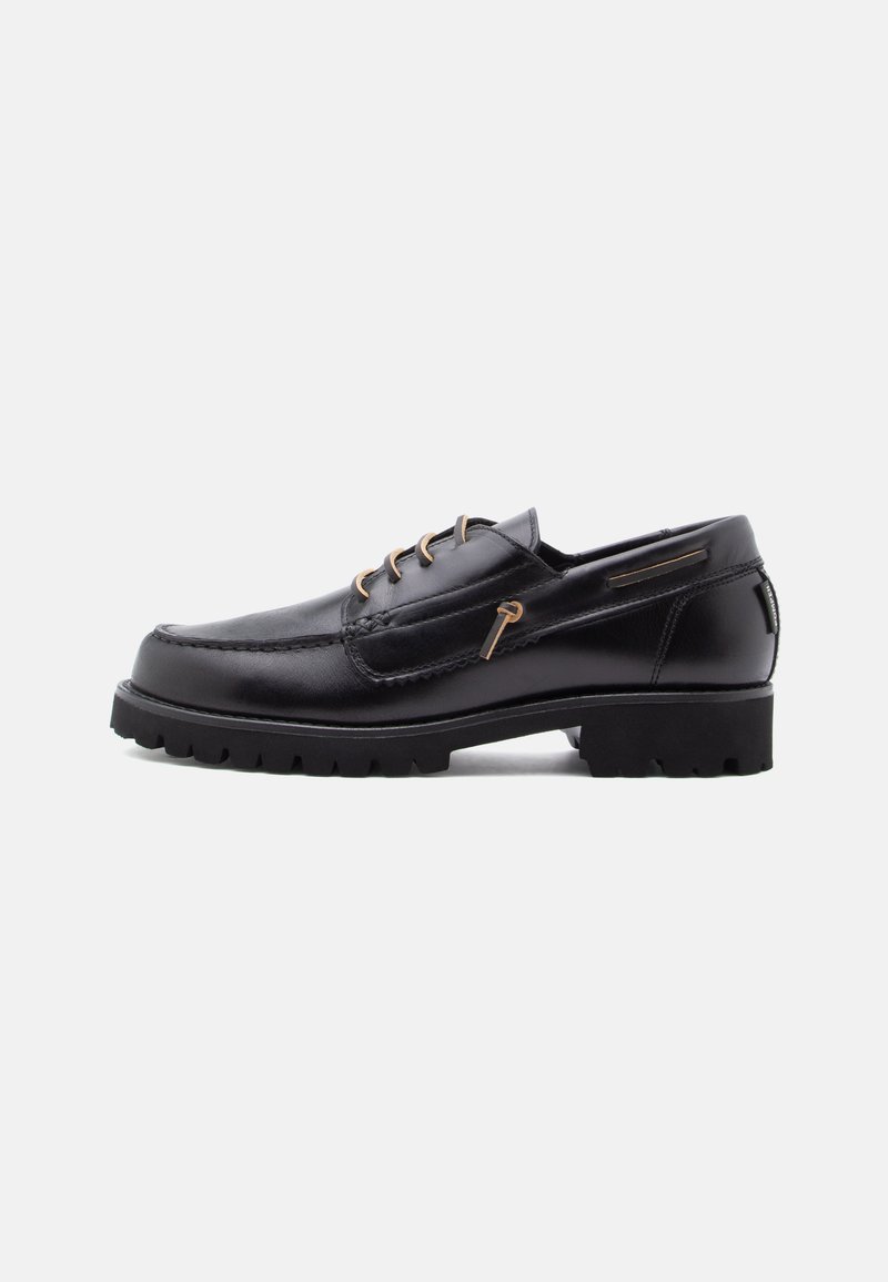 Chaussure en cuir noir avec un bout arrondi, quatre œillets marron, des coutures décoratives et une semelle en caoutchouc épaisse et texturée.