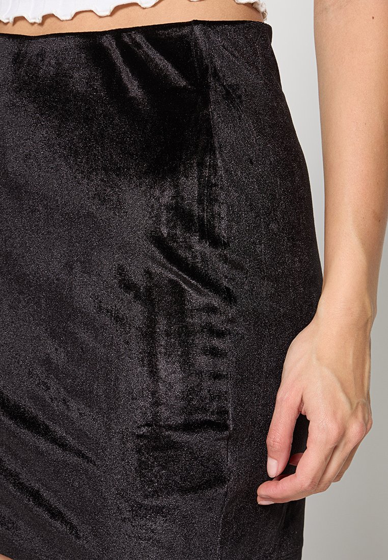 Czarna aksamitna mini spódnica z gładką strukturą, dopasowanym krojem, subtelnym połyskiem i z boku zamkiem błyskawicznym. Ręka spoczywająca na spódnicy.