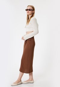 Bruine, gestructureerde midi-rok gecombineerd met een cropped witte trui met een ribbelpatroon. Geaccentueerd met zwarte zonnebril en lichte flats.