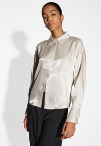 Camicia di satin beige chiaro con colletto e chiusura a bottoni, caratterizzata da maniche lunghe e una texture liscia e lucida. Indossata con pantaloni neri.