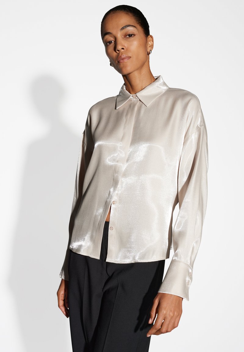 Camicia di satin beige chiaro con colletto e chiusura a bottoni, caratterizzata da maniche lunghe e una texture liscia e lucida. Indossata con pantaloni neri.