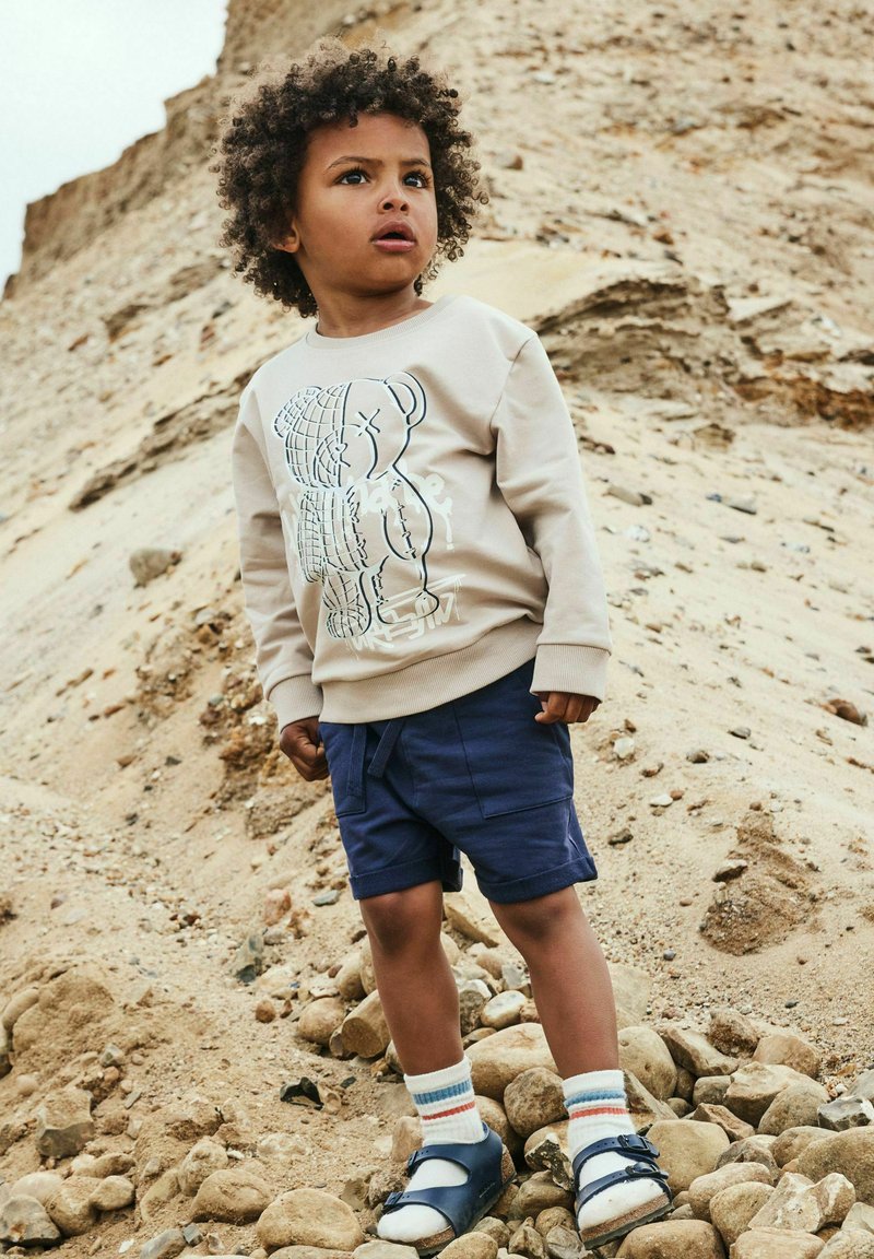 Jeune enfant avec des cheveux bouclés portant un sweat beige, un short navy, des chaussettes blanches et des sandales bleues, se tenant sur un terrain rocheux et sablonneux.