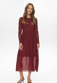 Rochie lungă burgundy cu mâneci transparente lungi, puncte texturate și un corp strâns care se transformă într-o fustă pliată. Pantofii cu toc negru completează look-ul.