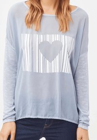Camiseta de manga larga azul claro con un corte relajado, que presenta un gráfico de corazón blanco y rayas verticales en el frente. Mangas suaves y texturizadas.