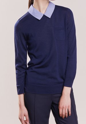 Person trägt einen marineblauen Pullover über einem blau-weiß gestreiften Hemd mit Kragen, kombiniert mit passenden marineblauen Hosen vor neutralem Hintergrund.
