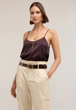 Femme portant un caraco en satin violet foncé rentré dans un pantalon crème taille haute avec une ceinture foncée et des bijoux en or, debout devant un fond uni.