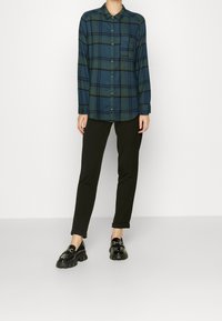 Zielono-niebieska koszula z długim rękawem, z kołnierzykiem i kieszenią z przodu, zestawiona z czarnymi spodniami podwiniętymi na dole oraz błyszczącymi czarnymi loafersami.