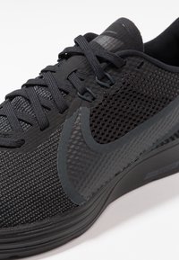 Baskets de sport noires avec un dessus en mesh texturé, des accents en plastique lisse, des lacets épais et une semelle rembourrée. Présente un logo proéminent dans une teinte plus foncée.