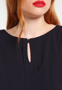Blusa azul marino con un escote redondo, que presenta un corte en forma de llave en la parte frontal, asegurado con un pequeño detalle de broche negro. Textura suave.