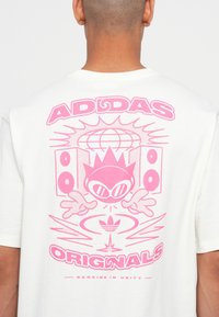 Dos d'une chemise blanche avec un graphique rose représentant un personnage de dessin animé, des haut-parleurs, un globe et le texte "ADIDAS ORIGINALS" et "DANCING IN UNITY."