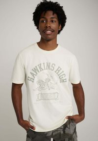 Bawełniany t-shirt w kolorze kremowym z napisem "HAWKINS HIGH" i ilustracją rodeo z tygrysem. Krótkie rękawy, luźny krój.