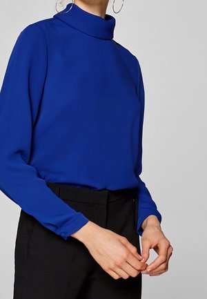 Blue high-neck blouse with long sleeves, smooth texture, fitted design, and button accents at the cuffs, paired with black trousers.