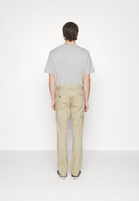 Man standing facing away wearing a light gray t-shirt, beige pants with a pocket on the side, and black slip-on shoes with white soles.