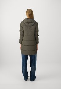 Manteau rembourré long, vert olive avec une capuche, présentant des sections matelassées horizontales et une coupe décontractée, associé à un jean bleu ample.