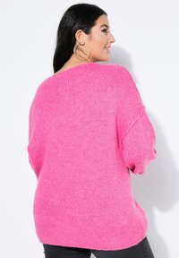 Oversized sweter w jasnoróżowym kolorze, o miękkiej, dzianinowej teksturze, z opuszczonymi ramionami i prążkowanymi mankietami. Nosi się go na czarnych spodniach.