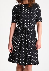 Czarna sukienka w białe kropki wykonana z miękkiego materiału, z okrągłym dekoltem, krótkimi rękawami oraz paskiem do związania w talii, rozkloszowanym dołem.