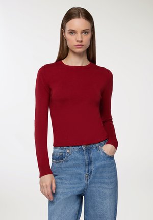 Maglione lungo rosso a maniche lunghe, dal design aderente, realizzato in tessuto morbido. Abbinato a jeans in denim azzurro chiaro a vita alta. Look minimalista, senza motivi.