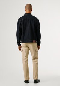 Chaqueta de mezclilla oscura con un corte recto, detalles de costura y una etiqueta de cuero marrón en la parte posterior, combinada con pantalones cargo color arena.