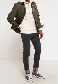 Olijfgroene jas met meerdere zakken over een crèmekleurige hoodie, gecombineerd met donkere jeans opgerold bij de manchetten en grijze sneakers met witte zolen.