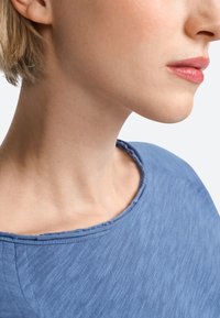 Kék, mintázott ruhadarab, széles, lekerekített nyakkivágással, egy személy nyakán és állvonala mentén látható, amely laza szabást és hétköznapi stílust mutat.