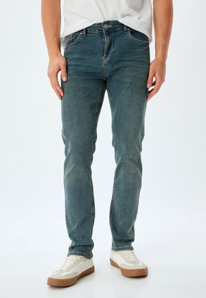 Jeans de mezclilla azul con corte ajustado, con cremallera, cinco bolsillos y un ligero desgaste. Combinados con zapatillas de deporte en color blanco roto.